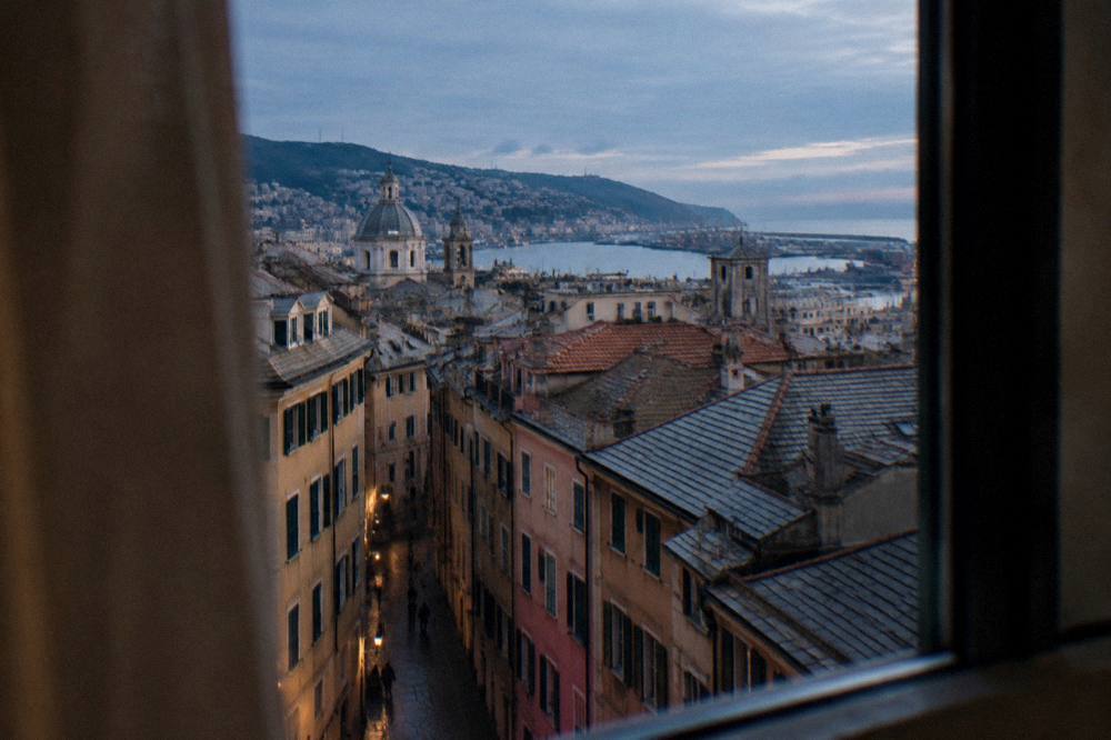 A hotel room window in Genoa looking out over tiled rooftops toward the harbour