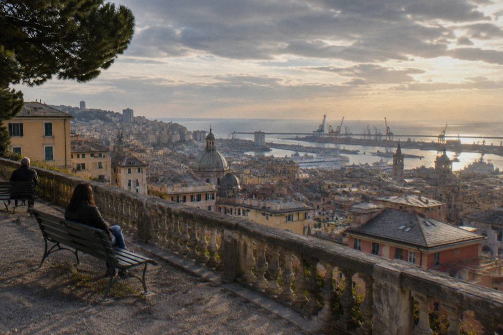 An open city terrace at dusk with rooftops descending toward a lit harbour