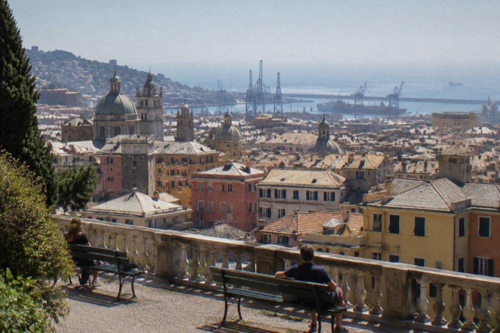 A hillside terrace with a stone balustrade overlooking terracotta rooftops, harbour cranes and a hazy sea