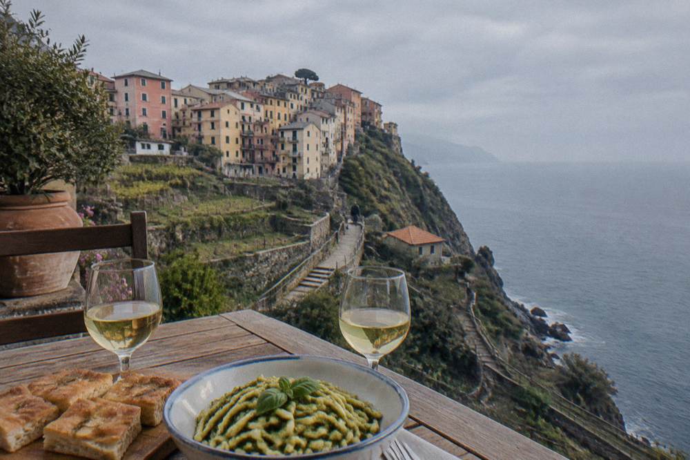 A table with focaccia, a bowl of trofie pasta with pesto and two glasses of white wine