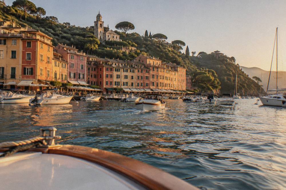 A boat approaching a small Mediterranean harbour lined with pastel buildings under a clear sky
